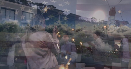Six friends holding sparklers beside patio chairs and low coffee tables at dusk, with string lights