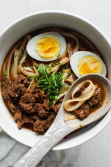 Overhead view of spicy noodle soup in a ceramic spoon, top view of noodles eggs in a spicy broth with scallions