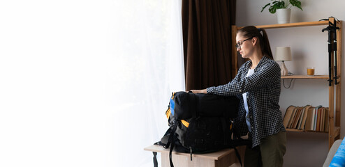 Preparing for a hike in the mountains. Young woman packing a backpack for a hiking trip in home interior. Banner.