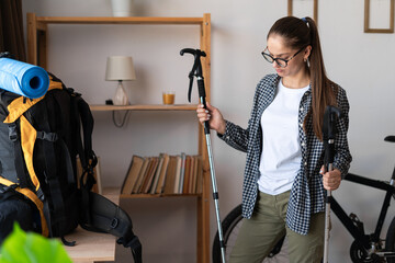 Young woman traveler hiker packing her backpack holding nordic poles ready for traveling at home...