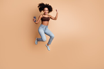 Excited young woman jumps with joy holding a smartphone on beige background, expressing confidence...