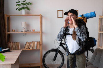 young female traveler hiker ready to trip with backpack screaming announcement at home, positive...