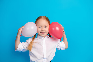 Smiling schoolgirl with braided red hair holding balloons against a blue background, showing...