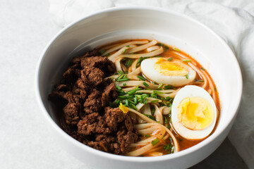 Overhead view of spicy noodle soup with eggs and beef chunks in a white bowl, top view of noodles eggs in a spicy broth with scallions