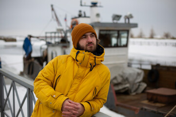 Travel photo. Young adult white man with beard, yellow winter jacket with hood, brown hat stands near fisher boat on sea shore, looks side. Cold winter weather. Tourism, trip, seafaring. Ship on coast