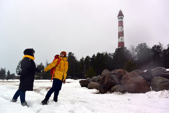 Horizontal travel photo. Young adult man with beard, yellow winter jacket, big red tourist backpack, mature woman are walking near Osinovetsky lighthouse. Ice, white snow on sea. Tourism, trip, walk
