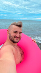 Vertical mobile selfie photo. Young adult tanned fat white man with mohawk, stubble, big belly, magenta swim ring on pebble sea beach looks at camera. Sun day. Blue sky. Travel, trip, summer tourism