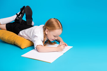 Smiling schoolgirl with red hair writing in a notebook against blue background, enjoying creative...