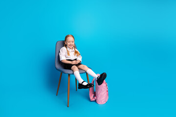 Smiling schoolgirl in stylish uniform sitting on chair with pink backpack in blue background...