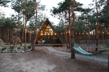 Modern A-frame cabin in a pine forest with hammock, trampoline, and string lights. Peaceful woodland retreat with cozy outdoor vibes