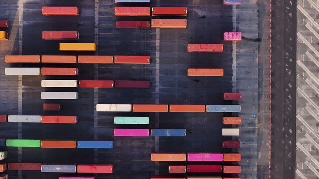 Port of Oakland, United States - 07 April 2025: Aerial view of a geometric tapestry of stacked shipping containers, their vibrant hues stark against the dark asphalt, creating an industrial mosaic.
