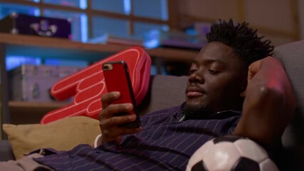 Side view shot of young Black man as sports fan watching sports match via smartphone while lying on sofa with football, copy space - Powered by Adobe