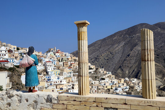 Statue of a Greek woman in traditional clothing overlooking the townscape of Olympos on the island of Karpathos, Greece, Greek columns with satutue at the entrance of the town olymbos