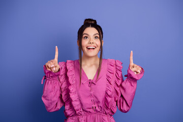 Adorable young woman in stylish pink dress pointing upward with happy expression in front of a...
