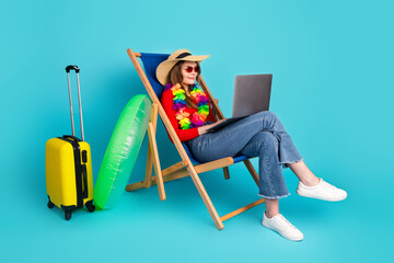 Cheerful young woman enjoying summer travel while working on a laptop, surrounded by vacation...