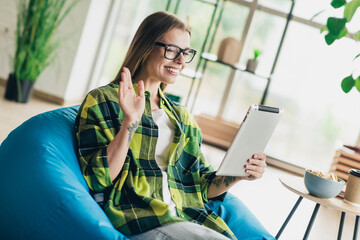 Positive woman waving on tablet while relaxing indoors during casual work or online study setting...