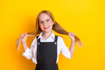 Smiling schoolgirl holding her hair strands wearing glasses and uniform against a bright yellow...