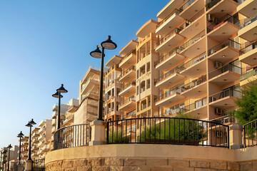 Low angle view of modern residential apartments with balconies along a curved stone promenade with black railings, street lamps, and green bushes under clear blue sky at sunset.