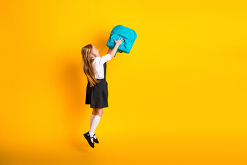 Smiling young girl in school uniform holding a blue backpack against a yellow background expressing...