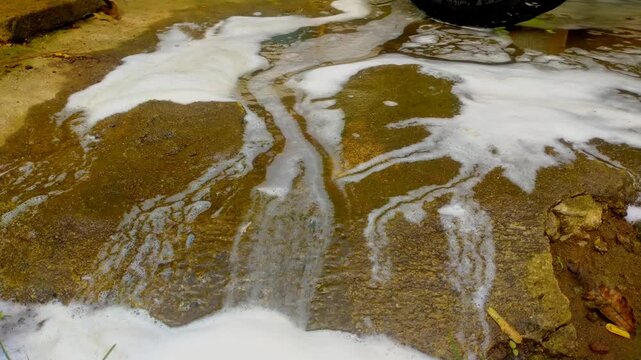 White, thick soap suds and murky water flow across a textured, dark concrete surface, with a small patch of green grass visible at the bottom.