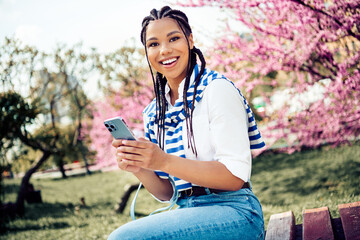 Cute woman sitting on a bench surrounded by blooming trees in a city park holding a smartphone and...