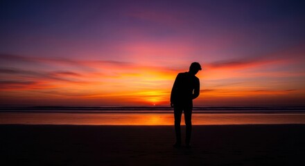 Silhouette of a person stands on a beach at sunset, the sky ablaze with vibrant colors, creating a serene and contemplative scene by the ocean.