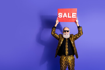 Stylish senior man in golden patterned suit holding sale sign against vibrant purple background...
