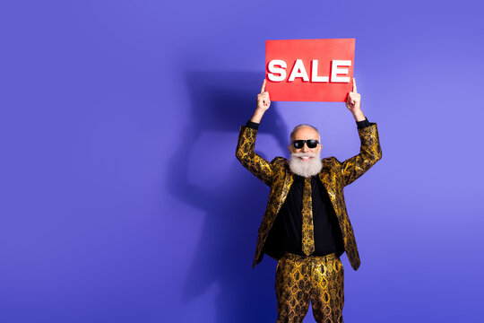 Stylish senior man in golden patterned suit holding sale sign against vibrant purple background showcasing luxury and extravaganza