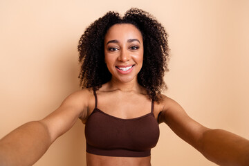 Young woman with curly black hair smiling at the camera in a brown top and beige background,...