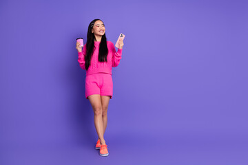 Smiling young asian woman in pink outfit holding a smartphone and cup on vibrant purple background
