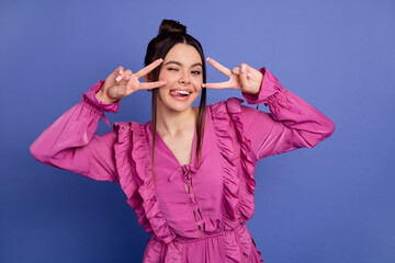 Joyful young woman in vibrant pink dress posing with playful peace sign on vivid purple background