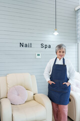 Senior Asian woman standing proudly wearing apron inside newly opened cozy nail spa boutique...
