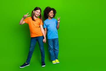 Two cheerful young friends holding hands making peace signs against vibrant green background