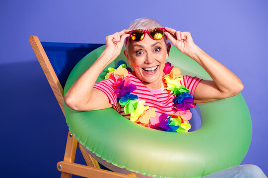 Cheerful elderly woman enjoying a summer vibe, wearing a colorful lei and sitting with a fun inflatable ring against a purple background.