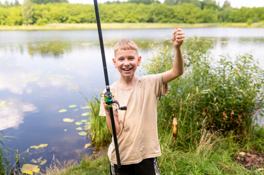 Happy teen boy holding fishing rod with caught fish on lake shore in summer