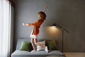 Cheerful young woman in a stylish orange shirt and white skirt dancing joyfully in a modern lounge...