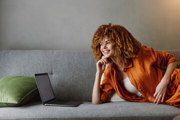 Happy young woman with curly hair relaxing on a gray sofa, wearing an orange shirt, smiling while...