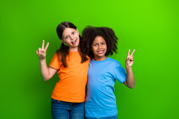 Two cheerful young girls posing on bright green background, showing peace hand gestures