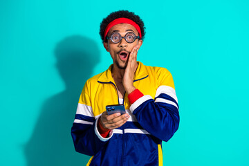 Excited young man in athletic retro outfit surprised while holding smartphone against turquoise background