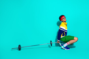 Young man in retro sportswear squatting with barbell against teal background showcasing vintage gym...