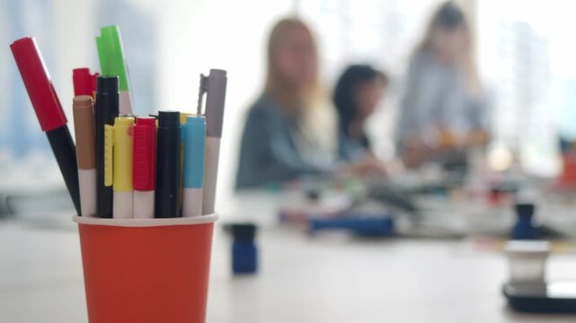 There are several multi-colored markers arranged in a cup, with a drawing workshop taking place in the background. Close up. Blur, defocused