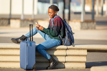Smiling African woman traveler using smartphone with backpack and suitcase