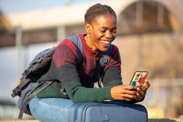 Happy African female traveler leaning on suitcase while texting outdoors