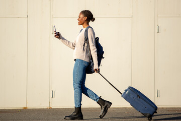 Young African woman pulling suitcase and checking phone while walking
