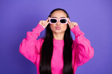 Cheerful young woman wearing pink sweater and glasses poses playfully in front of a solid purple...