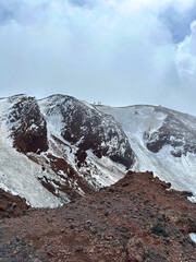 Snow-capped volcanic mountain landscape with rocky terrain, showcasing hikers on the summit, surrounded by dramatic clouds and a breathtaking natural environment