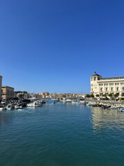 Fototapeta premium Scenic waterfront view showcasing boats moored along a tranquil harbor, with clear blue skies and historic architecture lining the shore, creating a picturesque coastal atmosphere