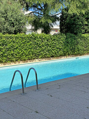 Swimming pool with clear blue water, surrounded by lush greenery and a paved area, featuring metal ladder for easy access, creating a serene outdoor relaxation space