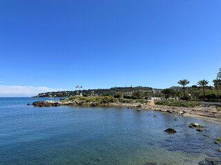 Serene coastal landscape featuring calm blue waters, rocky shoreline, and lush palm trees under a...