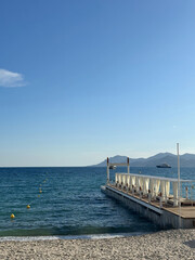 Fototapeta premium Serene beach scene with a wooden pier extending into calm blue waters, surrounded by gentle waves and distant mountains under a clear sky, evoking tranquility and relaxation